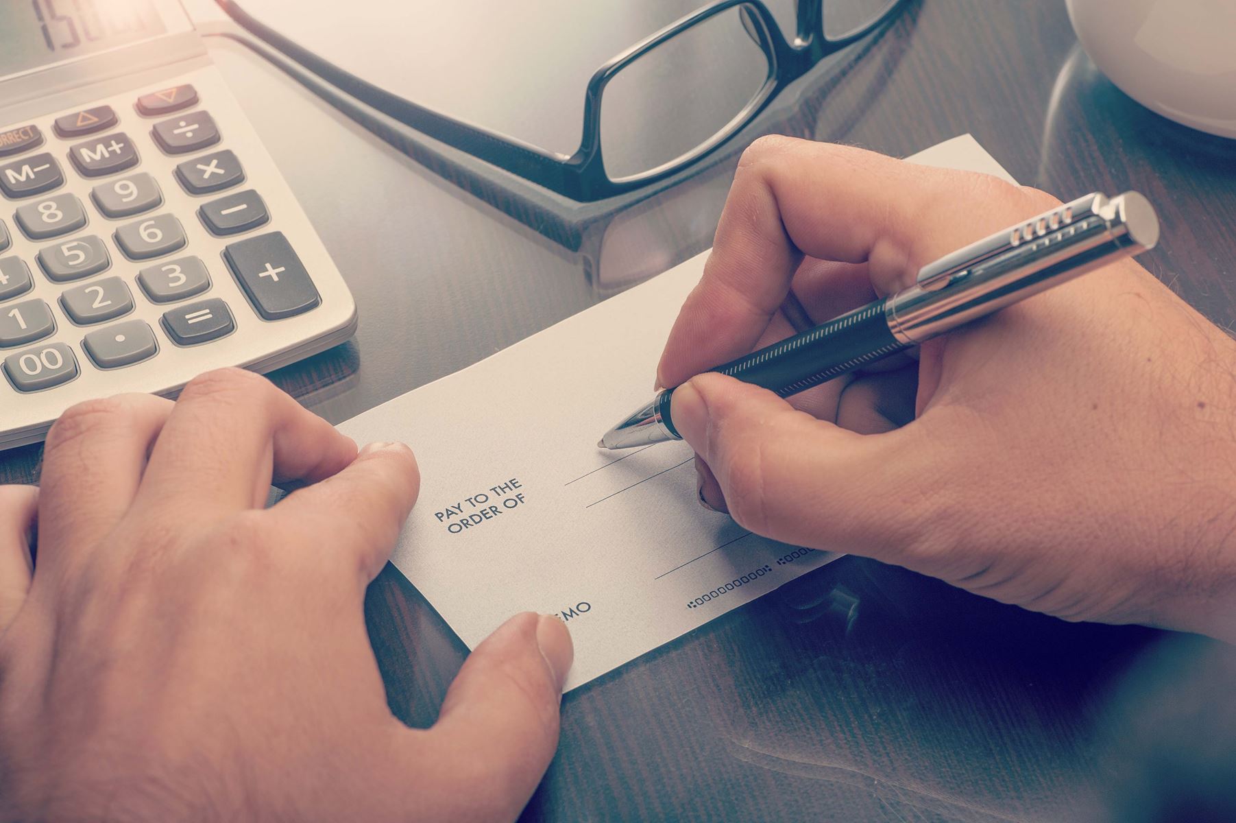 View of hands holding and writing a check, there is a calculator and a pair of glasses on the table too.