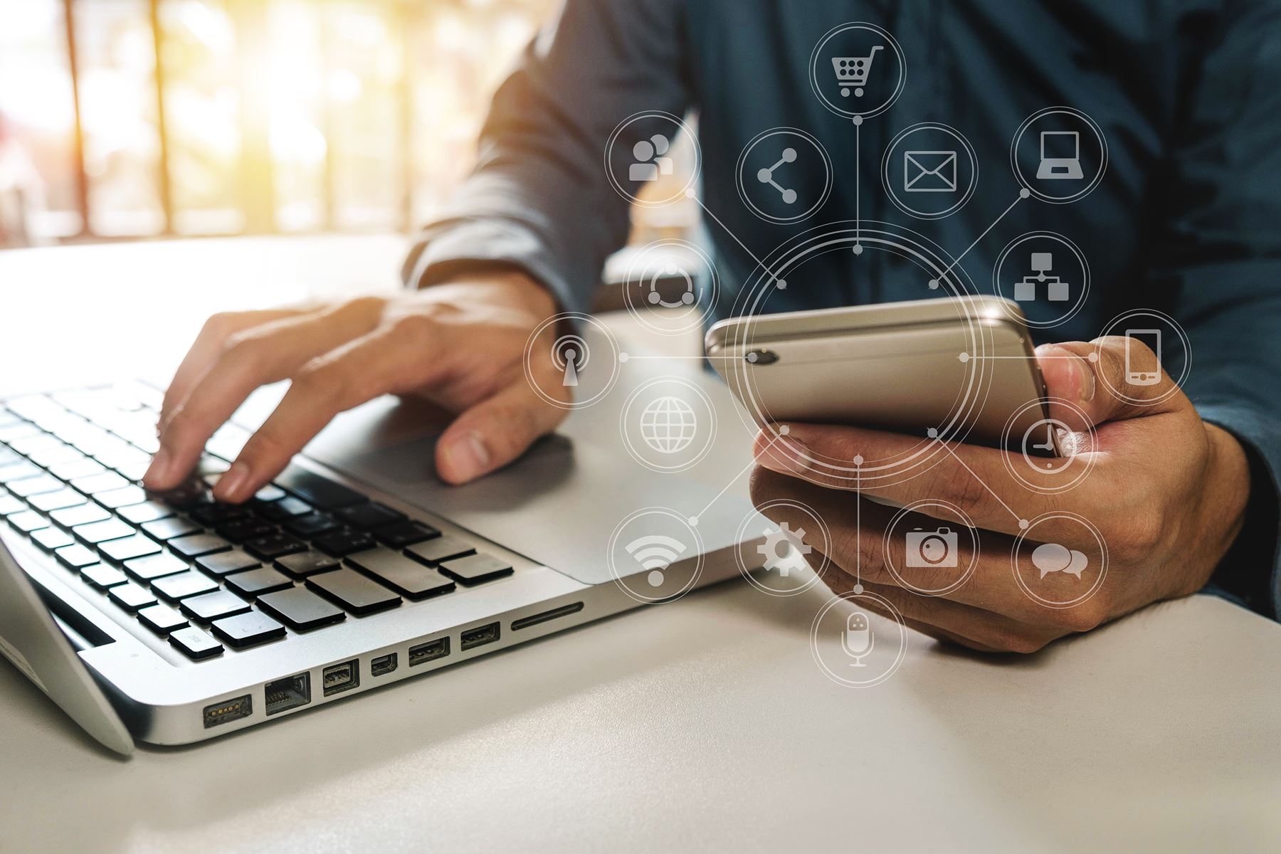 Man working with one hand on a laptop keyboard and the other hand is holding a cell phone